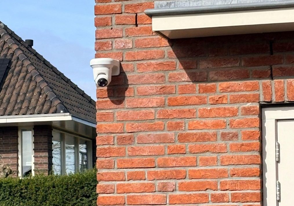 Witte camera op de hoek van een rode bakstenen gevel, met daken en een haag op de achtergrond